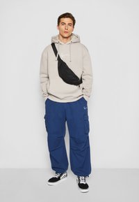 Beige hettegenser med logo, marineblå cargo-bukser med lommer, og svart bumbag. Svart og hvit sjakkmønstrede sneakers fullfører antrekket.