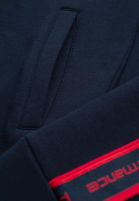 Marineblaue Sweatshirt mit glatter Textur, ausgestattet mit einer Vordertasche und roten Branding-Details entlang des unteren Randes.