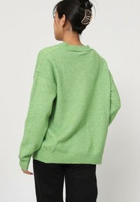 Pull en tricot vert avec une coupe décontractée, présentant un col et des poignets côtelés, vu de l'arrière, associé à un pantalon noir.