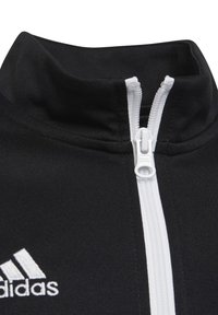 Schwarze Zip-Jacke mit hohem Kragen, ausgestattet mit einem weißen Reißverschluss und Akzenten. Beinhaltet ein weißes Logo auf der linken Brustseite. Glatte Stoffoberfläche.
