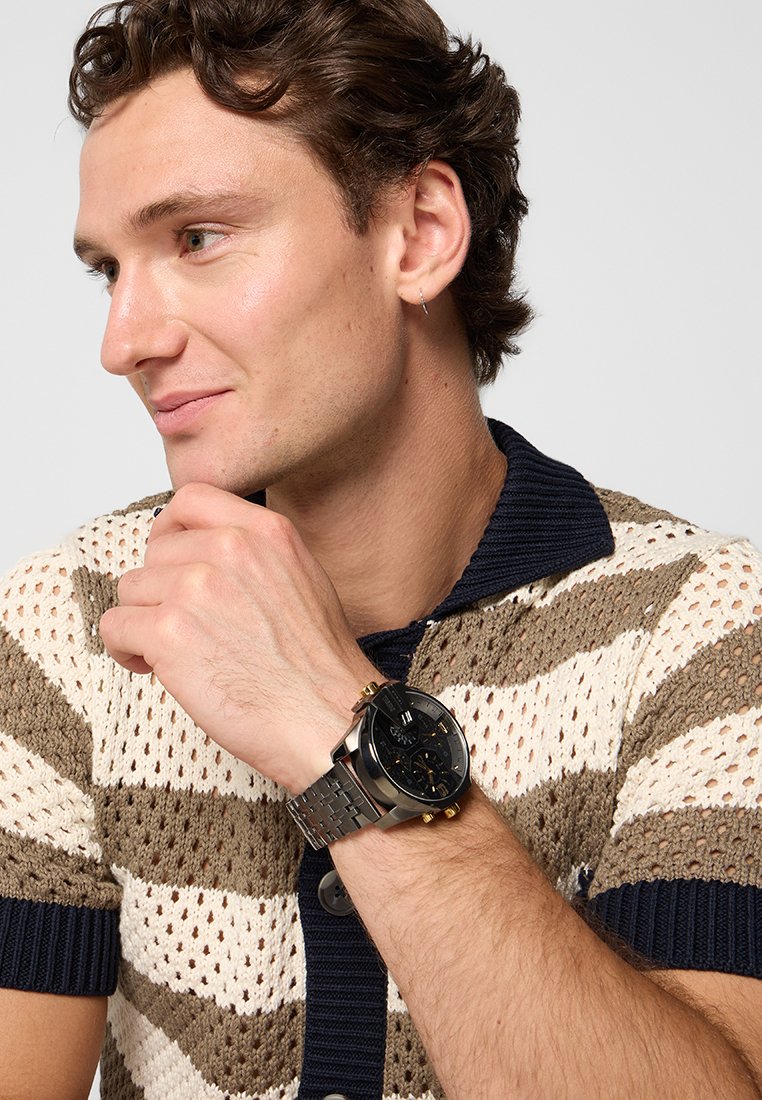 Montre noire avec bracelet en acier inoxydable, accents dorés, cadran rond et trois sous-cadrans. Chemise en tricot rayée brune et blanche texturée.