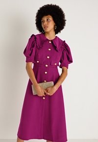 Vestido magenta com mangas bufantes, um colarinho franzido e botões dourados. Tecido suave com dois bolsos frontais. O modelo segura uma bolsa de mão.