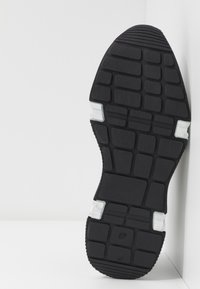 Semelle de chaussure en caoutchouc noir avec un motif de grille texturé, rehaussé de sections blanches. Présente un talon arrondi et une construction flexible.