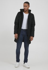 Parka negra con capucha sobre una camiseta blanca, combinada con jeans azul oscuro y zapatillas blancas. Tejido suave con un corte regular.