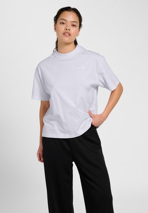 Jonge vrouw die een losse witte t-shirt en zwarte broek draagt, poseert met één hand in haar zak tegen een eenvoudige lichte achtergrond.