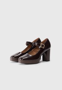 Brown patent leather Mary Jane heels with a rounded toe, ankle strap, and chunky striped wooden heel. Smooth texture and glossy finish.