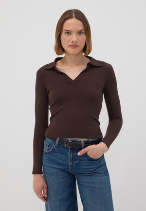 Mujer con el cabello castaño liso que lleva una camiseta de manga larga marrón oscuro y vaqueros azules, de pie con una mano en el bolsillo frente a un fondo liso.