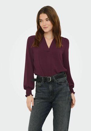 Femme aux longs cheveux bruns portant un chemisier prune à col en V avec manches bouffantes et un jean gris foncé avec une ceinture noire, debout devant un fond uni.