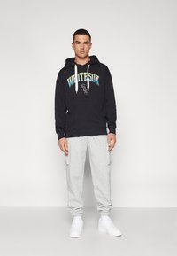 Sort hættetrøje med "WHITE SOX" logo, hvide snore, grå sweatpants med sidelommer, hvide sneakers, stående på en neutral baggrund.