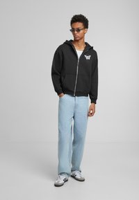 Sort zip-up hoodie med sommerfugle grafik; blanding af bomuld. Matchet med lyseblå jeans og hvide sko med sorte striber. Flad, afslappet pasform.
