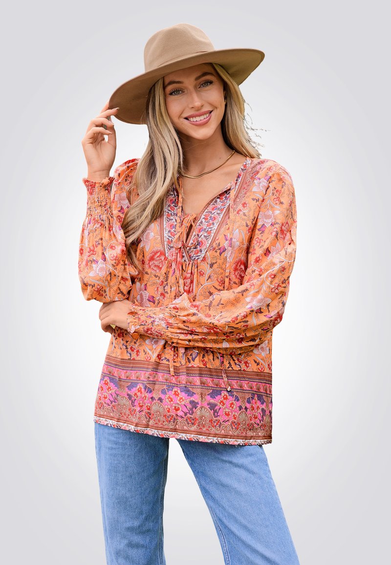Fließende orange Bluse mit Blumenmustern und elastischen Bündchen, kombiniert mit hellblauen Jeans. Das Modell trägt einen beige Hut mit breiter Krempe.