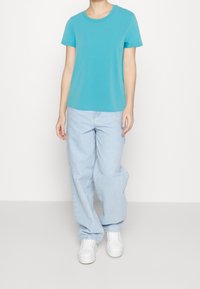 T-shirt turchese a maniche corte, colletto rotondo, abbinato a jeans azzurri larghi e sneakers bianche. Outfit casual e confortevole.