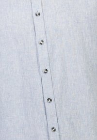 Camicia di lino azzurro chiaro con un tessuto texturizzato, caratterizzata da bottoni argentati lungo la patta frontale e dettagli di cucitura verticale.