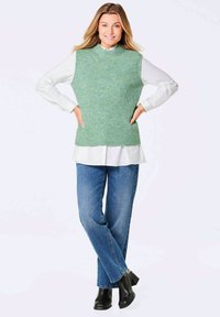 Groene gebreide mouwloze trui met een hoge hals over een wit shirt, gecombineerd met blauwe jeans en zwarte enkellaarsjes. Soepele textuur en een ontspannen pasvorm.