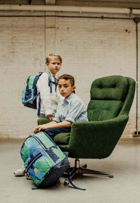Groene, gestructureerde fauteuil met chroombasis; twee kinderen met blauwe en groene rugzakken. Een jongen zit, de ander staat erachter, beiden in casual kleding.