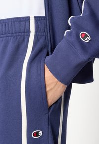 Μπλε σκούρο φούτερ με λευκό φερμουάρ, πλαϊνές τσέπες και λογότυπο Champion στο μανίκι. Το ύφασμα έχει ριμπ υφή και ρίγες.