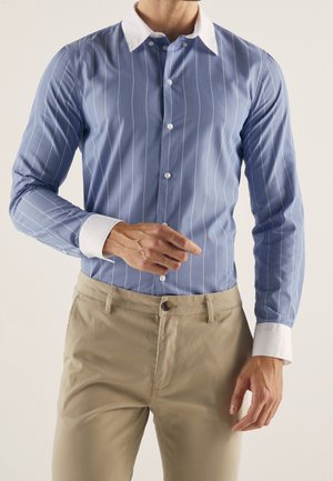 Mann trägt blau gestreiftes Langarmhemd mit weißem Kragen und Manschetten, kombiniert mit beigen Hosen, steht vor einem schlichten Hintergrund.