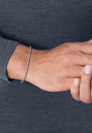 Bracelet en argent au design mince et flexible, avec une surface texturée et une finition lisse, porté sur un poignet avec un pull gris.