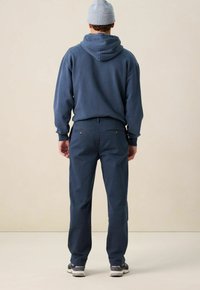 Blauwe hoodie met een ruime capuchon, gecombineerd met donkerblauwe broek. Bevat achterzakken met knopen en grijze sneakers.