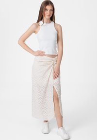 Witte geribde tanktop gecombineerd met een crèmekleurige maxi rok met een geometrisch kantpatroon en een zijsplit. Model draagt witte sneakers.