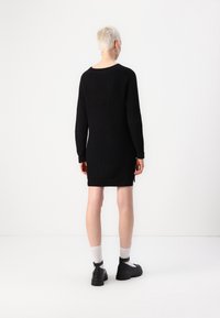 Personne avec les cheveux blonds courts portant une robe en tricot noire à manches longues, des chaussettes blanches et des chaussures noires épaisses, se tenant contre un fond blanc.