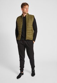 Gilet verde oliva imbottito con colletto alto, design trapuntato e chiusura con zip, indossato sopra una maglietta nera a maniche lunghe e pantaloni jogger neri con polsini elasticizzati.