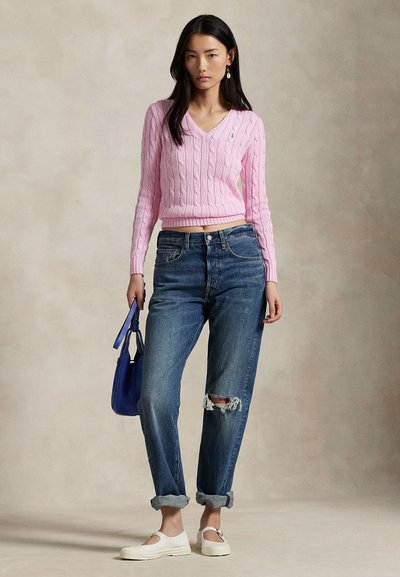 Junge Frau trägt einen rosa Pullover mit Zopfmuster, zerrissene hochgekrempelte blaue Jeans, weiße Schuhe und hält eine blaue Handtasche vor neutralem Hintergrund.