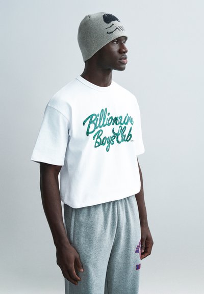 T-shirt bianca in cotone con logo "Billionaire Boys Club" verde in paillettes, abbinata a pantaloni della tuta grigi in cotone con scritta viola. Berretto grigio a maglia.