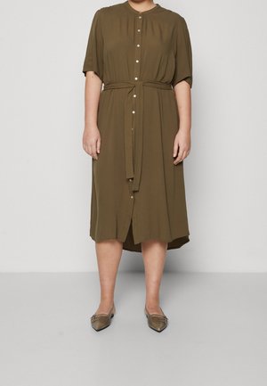 Person trägt olivgrünes, knopfgeschlossenes Midikleid mit kurzen Ärmeln und gebundenem Taillengürtel, steht in taupefarbenen spitzen Ballerinas auf grauem Boden.