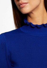 Primer plano de una mujer que lleva un suéter acanalado azul real con un cuello de mock rizado, mostrando parte de su cara y cuello.