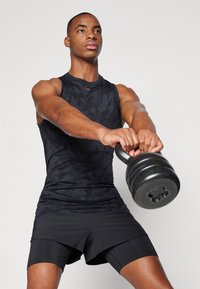 Svart ermeles t-skjorte med subtilt kamuflasjemønster, iført svarte shorts, holder en svart kettlebell.