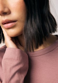 Långärmad tröja med rund hals i mjukt, mauve-färgat tyg. Närbild visar en slät textur och varsam sömnadsdetalj längs kragen och muddarna.