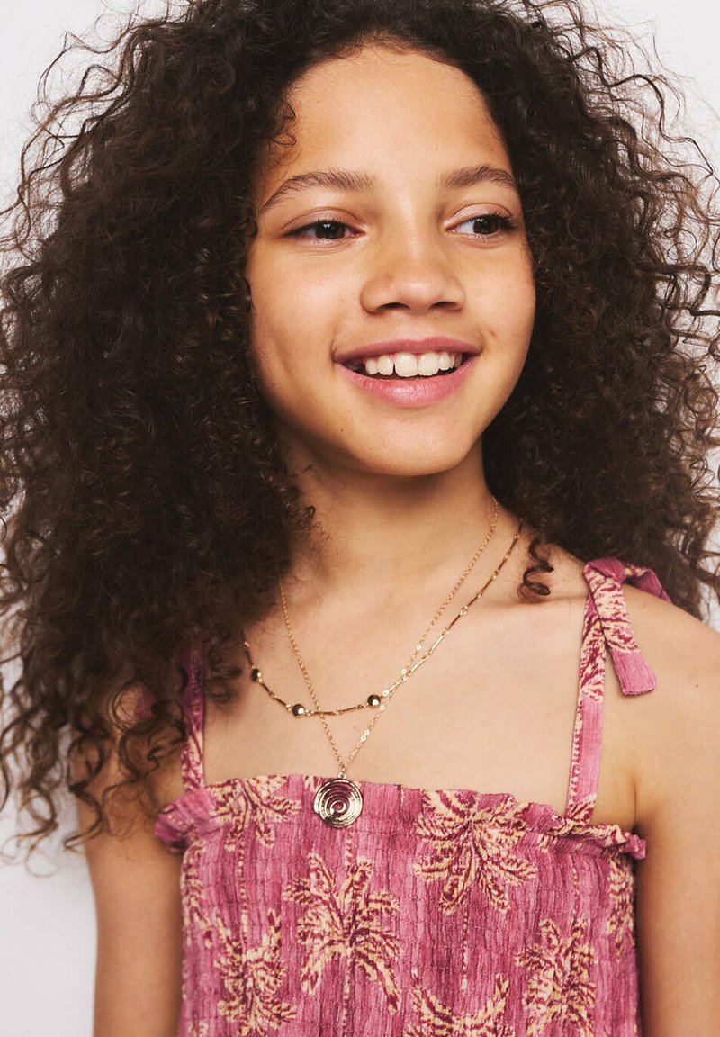 Fille souriante aux cheveux bouclés portant un haut sans manches rose à fleurs et des colliers en or superposés sur un fond uni.