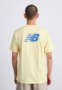 Geltona medvilninė marškinė, kurių gale yra didelis mėlynas "NB" logotipas. Trumpomis rankovėmis ir laisvu siluetu, minkštas audinys.