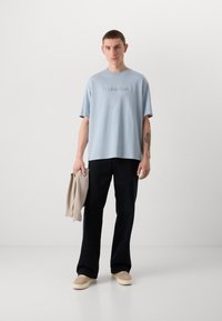 Light blue cotton t-shirt with short sleeves and embroidered brand logo. Paired with dark pants and holding a beige sweater, standing on a white backdrop.