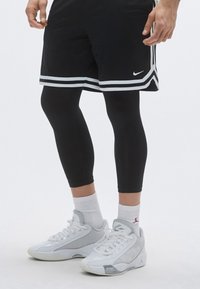 Μαύρο αθλητικό σορτς με λευκές ρίγες, μαύρα συμπιεστικά κολάν, άσπρες κάλτσες και λευκά-γκρι αθλητικά παπούτσια με λεπτομέρειες από δίχτυ.