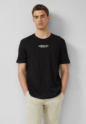 Jonge man met de handen in de zakken, gekleed in een zwart "s.Oliver Co." T-shirt en beige broek, tegen een effen achtergrond.