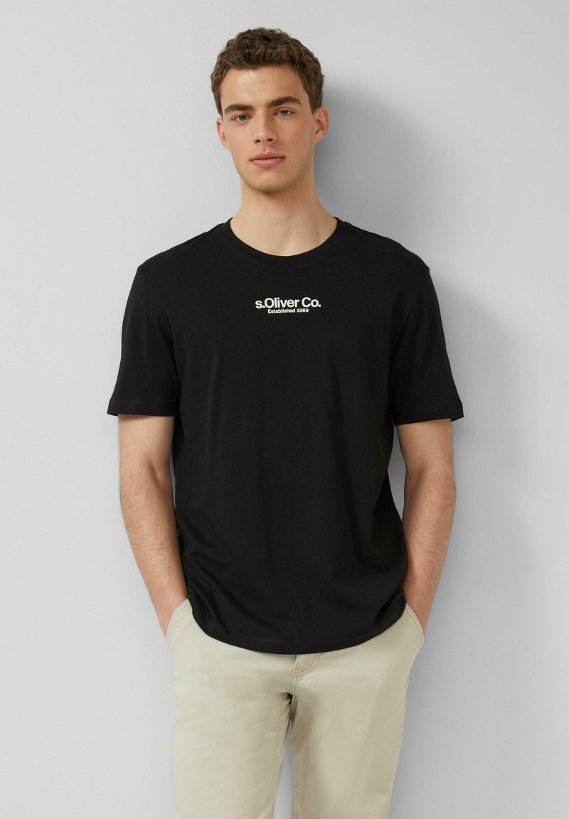 Jeune homme debout, les mains dans les poches, portant un t-shirt noir "s.Oliver Co." et un pantalon beige sur un fond uni.