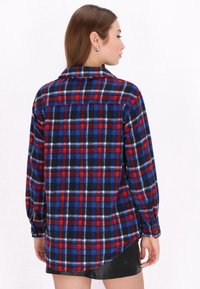 Camicia di flanella rossa, blu e bianca con un motivo a quadri, maniche lunghe e orlo curvo. Presenta un colletto e polsini con bottoni.