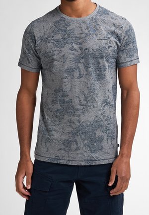 Mand iført en grå T-shirt med korte ærmer med et mørkt, abstrakt blomsterprint og mørke cargobukser.