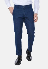 Pantaloni in denim blu scuro con vestibilità slim, dotati di chiusura a bottone, tasche laterali e una texture liscia, abbinati a scarpe nere lucide.