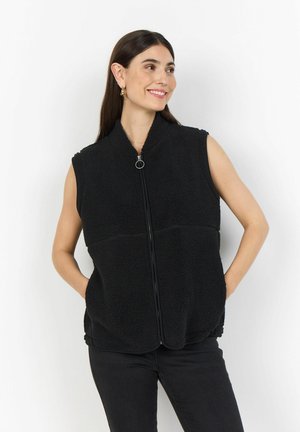 Woman smiling and looking sideways wearing sleeveless black fleece vest with front zipper and hands in pockets.