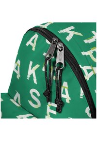 Mochila verde con grandes letras blancas y acentos amarillos. Cremallera negra con tiradores de metal y cordón texturizado. Superficie de tela suave.