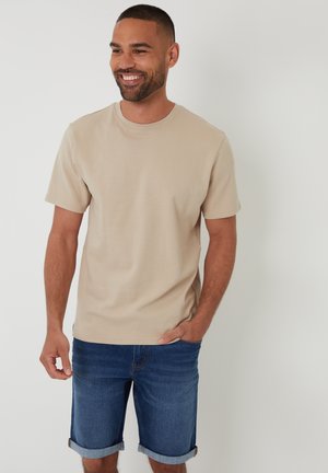 Beige T-Shirt mit kurzen Ärmeln und Rundhalsausschnitt, aus strukturiertem Stoff, kombiniert mit hochgekrempelten blauen Jeansshorts. Lässige Passform.