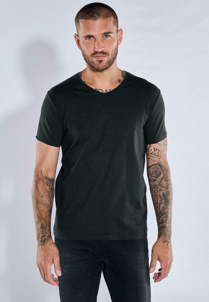 Schwarzes V-Ausschnitt-T-Shirt aus glattem Baumwollmaterial; kurze Ärmel; getragen mit dunklen Jeans; zeigt sichtbar tätowierte Arme als markantes Detail.