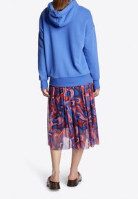 Sudadera azul con un ajuste relajado, combinada con una falda fluida y semitransparente con patrones de remolinos en rojo y rosa. Sandalias plataforma marrones.