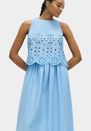 Vrouw draagt lichtblauwe mouwloze jurk met bloemenuitgesneden patronen op het lijfje, handen rusten achter haar rug, kijkt naar beneden.