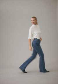 Valge lühikeste varrukatega pluus koos hõlpsalt laienevate siniste teksapükstega. Sile tekstuur ja klassikaline siluett. Modell liikumises halli tausta ees.