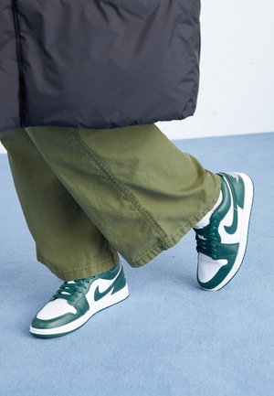 Person iført grønne og hvide sneakers og løstsiddende olivengrønne bukser, stående på lyseblåt tæppe.