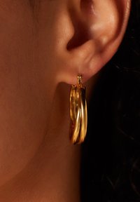 Gouden hoepel oorbellen met een gedraaide design en een gladde afwerking, die een weerspiegelende textuur tonen en een kleine studdetails aan de bovenkant hebben.
